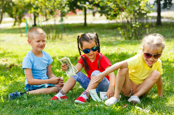 Twee kleine meisjes en jongen met stapel dollars zittend in groen gras in Park - Foto, afbeelding