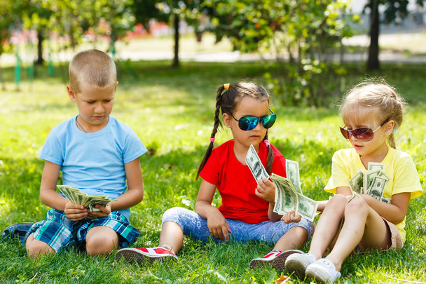 Deux petites filles et un garçon avec une pile de dollars assis dans l'herbe verte dans le parc
 - Photo, image