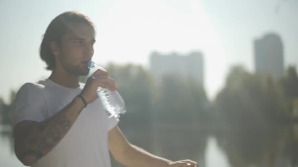 Handsome male drinks water from bottle in the park - Materiał filmowy, wideo