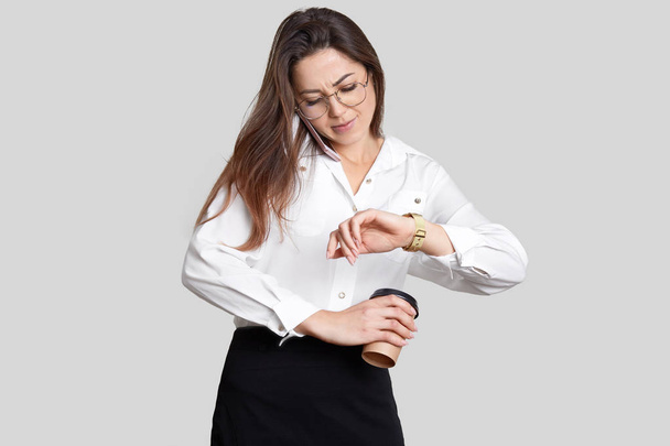 Busy young female has telephone conversation, hurries at work, looks at watch, holds aromatic takeaway coffee, dressed in formal clothes, isolated over white background. Business and work concept - Foto, Imagen