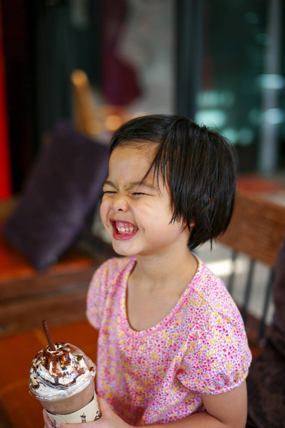Portriat азіатські маленька дівчинка пити змішані лід шоколадом з смішне обличчя. Щаслива дівчина насолоджуватися їжею безалкогольний напій з великий усмішкою і ямочки на щоці в кафе - Фото, зображення