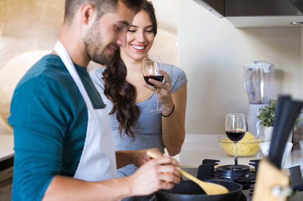 Shot of beautiful young smiling woman drinking red wine while her husband preparing lunch in the kitchen at home. - Foto, imagen