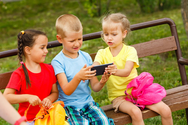 Tři šťastné děti hrají s smartphone na lavičce v parku  - Fotografie, Obrázek