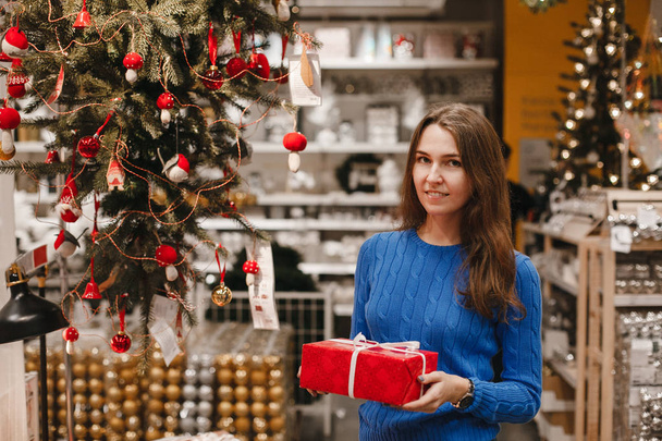 Happy woman with red present gift box near Christmas tree decoration in New Year shop. Brunette smiling girl at christmas market. Shopping. Lifestyle. - Fotografie, Obrázek