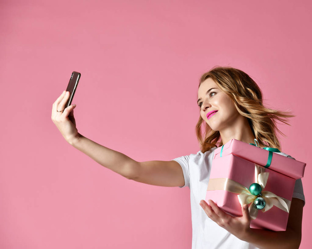 若くてきれいな女性保持クリスマス贈り物笑顔と彼女の携帯電話でモバイル selfie 写真を作る - 写真・画像