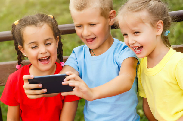 Tres niños felices jugando con teléfono inteligente en el banco en el parque
  - Foto, imagen