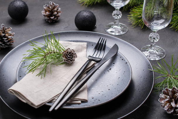 Темно-різдвяний дизайн столу. Чорні тарілки, келихи для шампанського, виделка та набір ножів з серветкою, ялинкова гілка, різдвяні прикраси
 - Фото, зображення