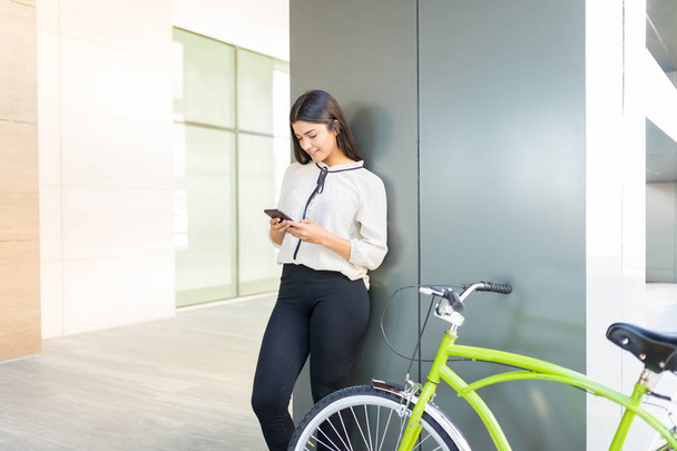 Empleada que utiliza aplicaciones móviles en bicicleta verde contra columna
 - Foto, Imagen