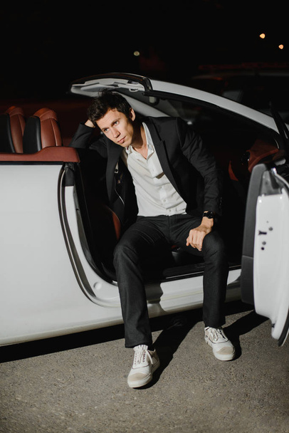 Stylish young man sitting posing in his white cabriolet. Nightlife. Businessman in suit in luxury car - Fotó, kép