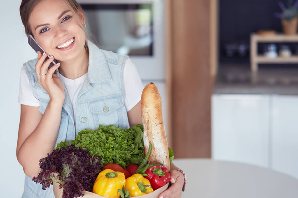 Mujer sonriente con teléfono móvil sosteniendo bolsa de compras en la cocina - Foto, Imagen