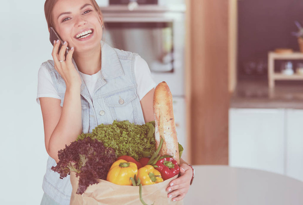 Smiling woman with mobile phone holding shopping bag in kitchen - Fotografie, Obrázek