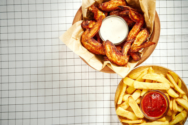 Damalı masa örtüsü üzerinde lezzetli kızarmış tavuk kanatları, patates kızartması ve soslar üstten görünüm - Fotoğraf, Görsel
