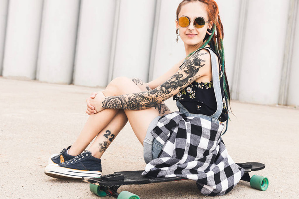 young girl with tattoo and dreadlocks sitting on a longboard on the background of concrete structure - Foto, Imagem
