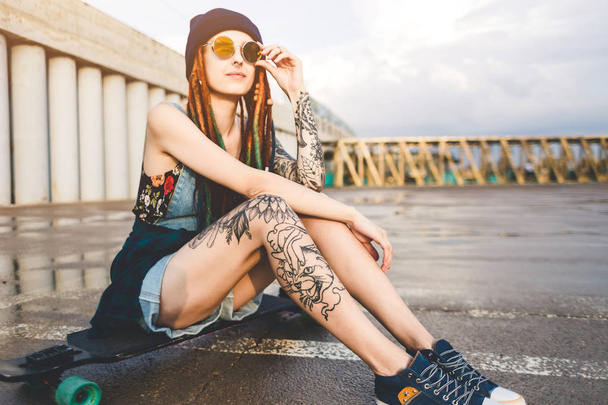 young girl with tattoos and dreadlocks in a blue cap sits on a longboard against the background of concrete structure - Photo, image