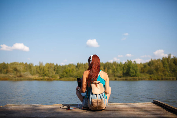 Photo on back of woman in headphones with backpack and laptop in hands sitting on river bank - Фото, изображение