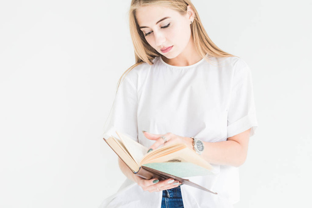 Portrait of a young stylish blonde girl in a white T-shirt and blue jeans reading a book on a white background - Photo, Image
