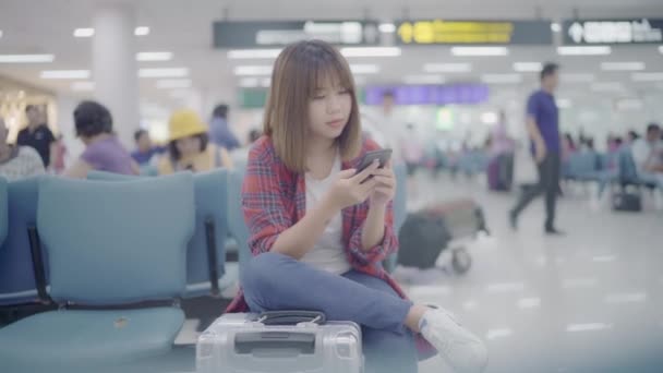 Szczęśliwy Asian kobieta przy użyciu i sprawdzanie jej smartphone, siedząc na krześle w hali terminala podczas oczekiwania jej lotu przy bramce w międzynarodowy port lotniczy. Kobieta szczęśliwa w koncepcji Lotnisko. - Materiał filmowy, wideo