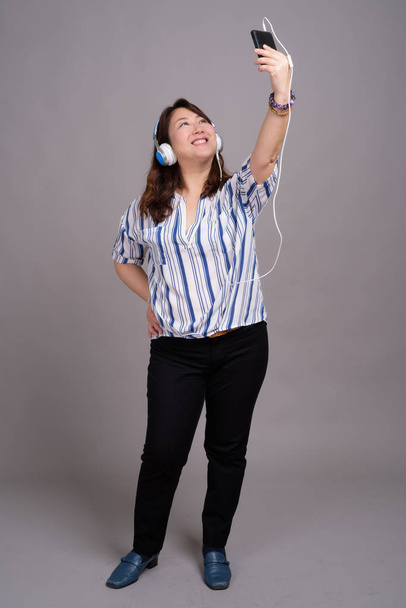 Portrait of mature Japanese businesswoman with headphones taking selfie - Photo, Image