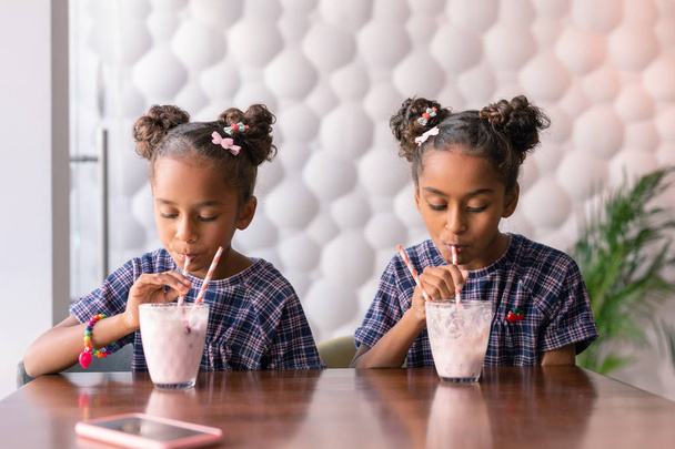 Dark-haired sisters wearing bright hair slides enjoying their milkshakes - Fotografie, Obrázek