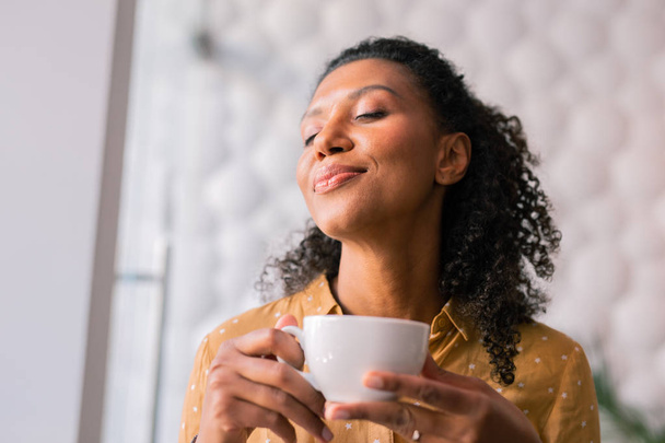 Curly dark-haired appealing woman feeling joyful while drinking coffee - Foto, Imagen
