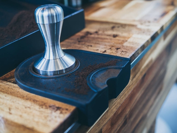 Stainless steel coffee tamper and ground coffee on wooden table in cafe. - Foto, Imagen