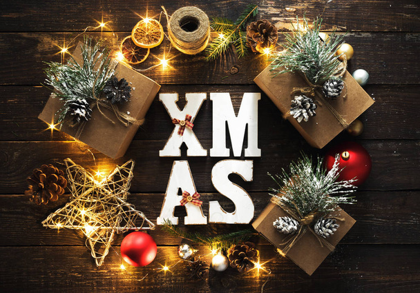 Flat lay Christmas background. Word Xmas and wreath made of Christmas decoration and christmas lights on dark wooden background top view - Foto, Imagem