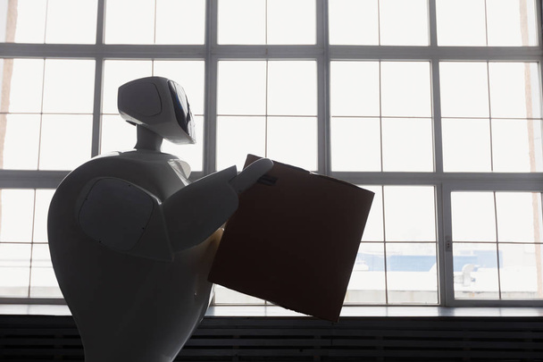 um robô com caixa de papelão na mão. conceito de entrega. silhueta do robô contra a janela
 - Foto, Imagem