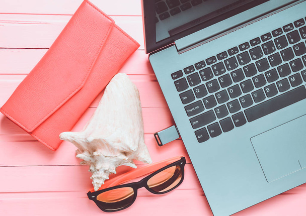 das Konzept der Web-Arbeit in einem Badeort, freiberuflich. Laptop, Flash-Laufwerk, Sonnenbrille, Handtasche auf einem rosafarbenen Holztisch. Ansicht von oben. - Foto, Bild