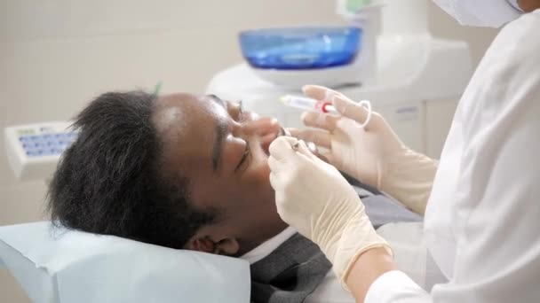 歯科医は、顎でノボカインを凍結のショットを置きます。歯科医院の椅子に男性患者は若いアフリカ系アメリカ人。医学、健康、口腔病学の概念. - 映像、動画