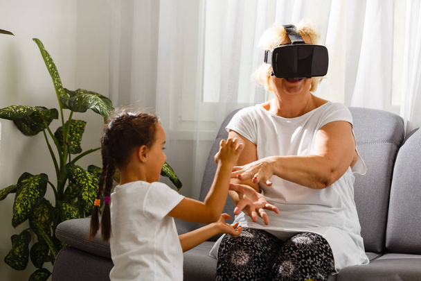 Старша жінка використовує окуляри віртуальної реальності з онукою вдома
 - Фото, зображення