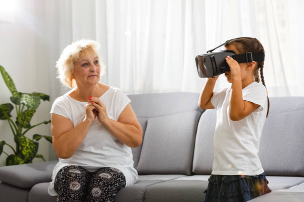Senior woman using virtual reality glasses with granddaughter at home - Foto, afbeelding