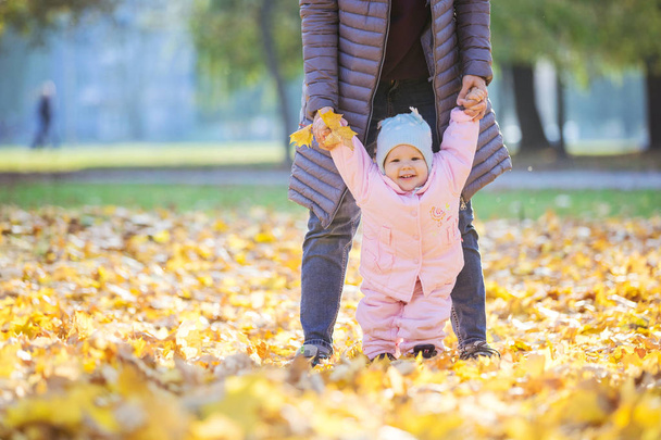 Mother supporting baby daughter and helping her make first steps. Mom and daughter on walk in autumn park. - Photo, Image