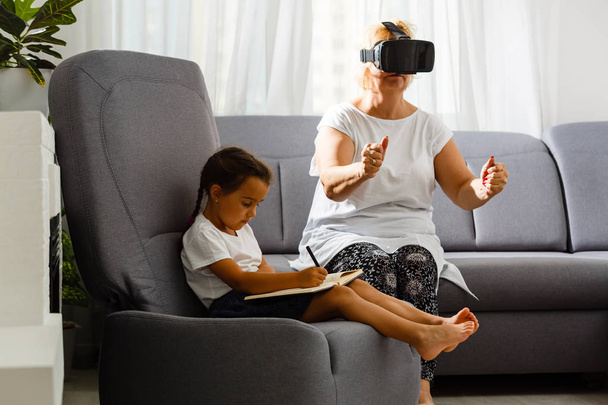 Ανώτερος γυναίκα χρησιμοποιώντας γυαλιά εικονικής πραγματικότητας με την εγγονή του στο σπίτι - Φωτογραφία, εικόνα