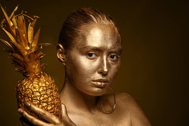 Красива молода жінка з золотою фарбою на її тілі і ананасом на темному фоні
 - Фото, зображення