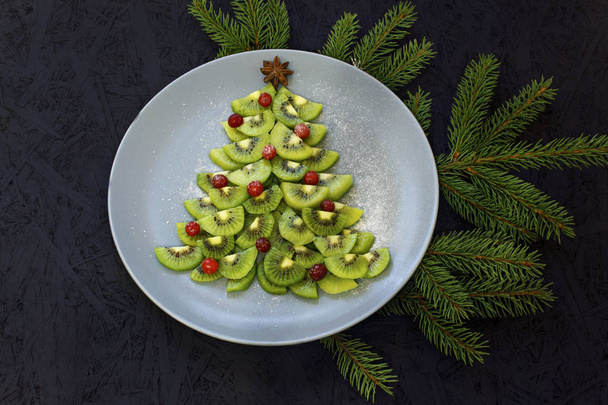 Kiwi-Scheiben auf einem Teller in Form eines Weihnachtsbaums auf schwarzem Hintergrund ausgebreitet. Blick von oben. Neues Jahr und Weihnachten. - Foto, Bild