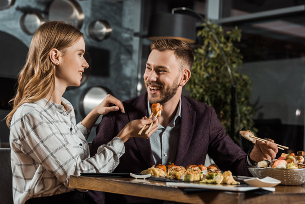 Beautiful smiling young woman feeding her boyfriend with sushi in restaurant - Foto, afbeelding