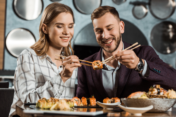 Smiling attractive young adult couple eating sushi together in restaurant - Photo, image
