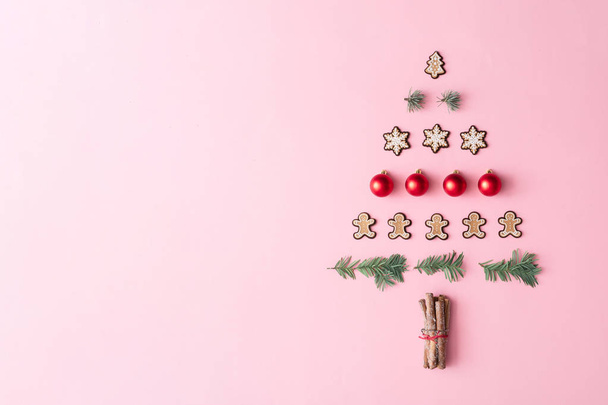 Christmas tree made with evergreen fir branches and bauble decorations with winter cookies and cinnamon sticks on pastel pink background. Minimal holiday flat lay table composition  - Photo, Image