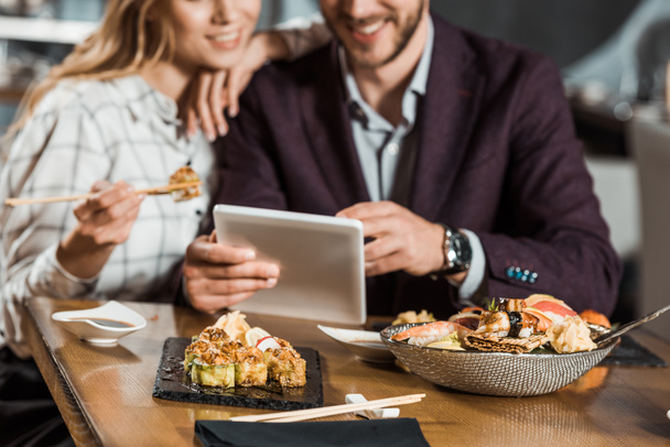 Rajattu näkymä pari käyttää digitaalista laitetta syödessään sushia ravintolassa
 - Valokuva, kuva
