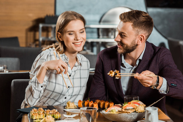 Attractive smiling couple eating together sushi rolls in restaurant  - Photo, Image