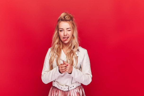 Young cheerful woman with long blonde wavy hair standing on red background and sticking out tongue. - 写真・画像