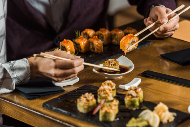 Vista recortada de la gente sumergiendo rollos de sushi en salsa de soja en el restaurante
 - Foto, Imagen