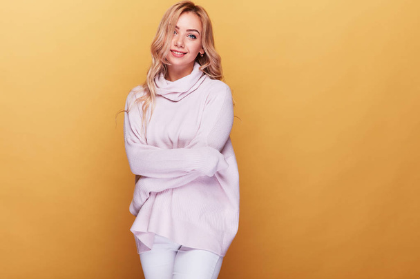Young cheerful woman with long blonde wavy hair and in light pink sweater standing on yellow background. - Φωτογραφία, εικόνα