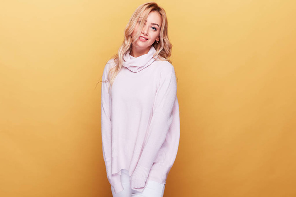 Young cheerful woman with long blonde wavy hair and in light pink sweater standing on yellow background. - Foto, Bild