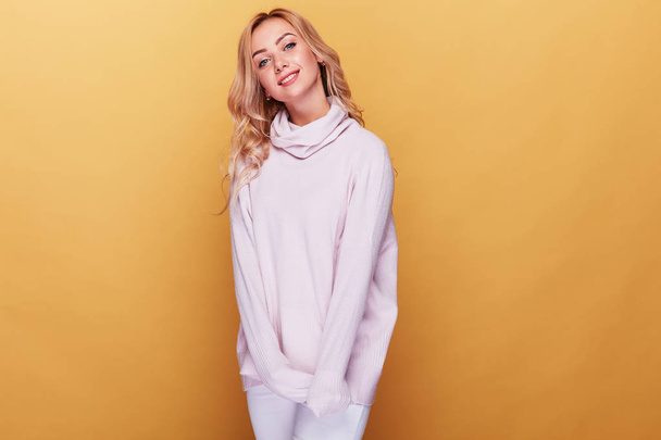 Cute woman with long blond wavy hair dressed in light pink sweater standing on yellow background  - Fotoğraf, Görsel