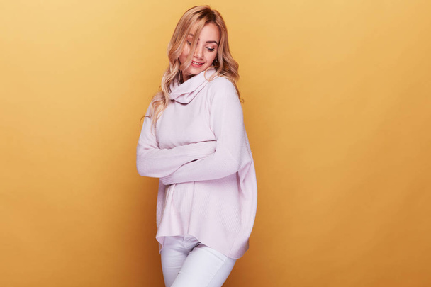 Cute woman with long blond wavy hair dressed in light pink sweater standing on yellow background with her eyes closed - 写真・画像