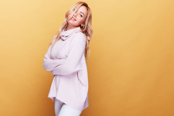 Cute happy woman with long blond wavy hair dressed in light pink sweater standing on yellow background  - Foto, afbeelding