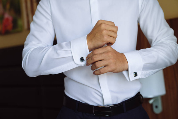Closeup επιχειρηματίας βάζει μανικετόκουμπα, φοράει ακριβό δερμάτινη ζώνη. Ο άνθρωπος σε ένα business κοστούμι, λευκό πουκάμισο. Προετοιμασία του γαμπρού την ημέρα του γάμου - Φωτογραφία, εικόνα
