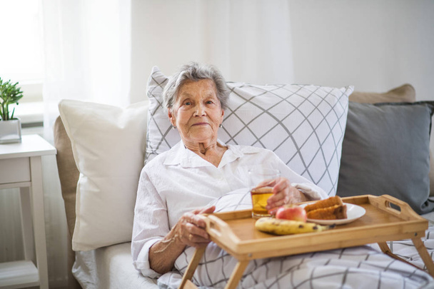 An elderly sick senior woman with food on a wooden tray lying in bed at home. - Valokuva, kuva