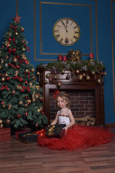 cute little girl at the Christmas tree in a red elegant dress with a box with gifts in her hands - Photo, image
